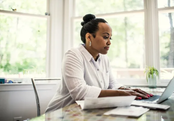 Woman typing on laptop at kitchen table near large windows